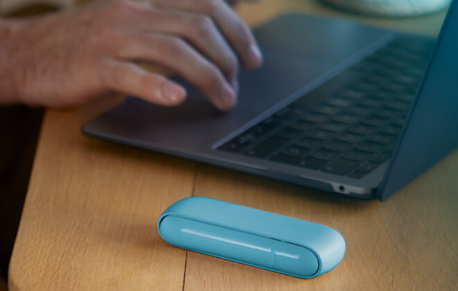 Hände tippen auf einem Laptop und einem iPhone. Daneben liegt ein IQOS ORIGINALS DUO Pocket Charger in türkiser Farbe.