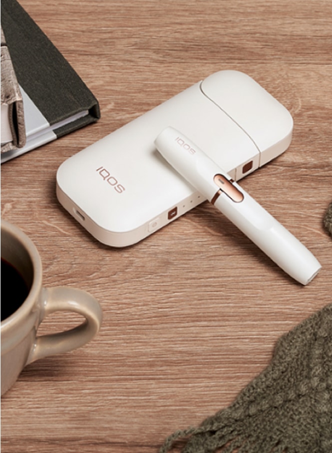 White IQOS 2.4 PLUS Holder and Pocket Charger on a wooden table next to a cup